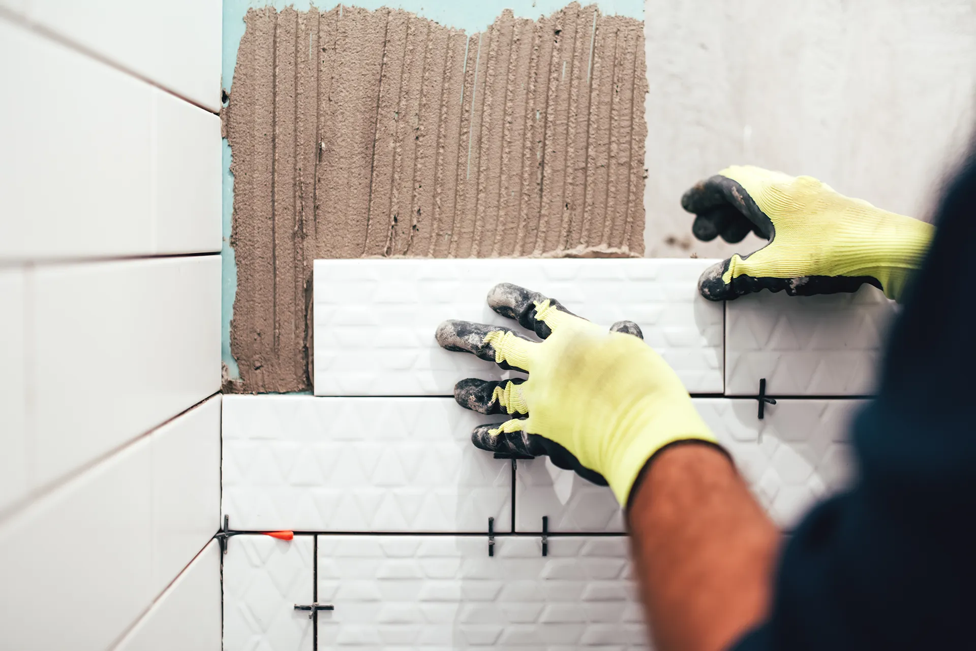 Worker wearing gloves installing white subway tile with adhesive and tile spacers on a bathroom wall