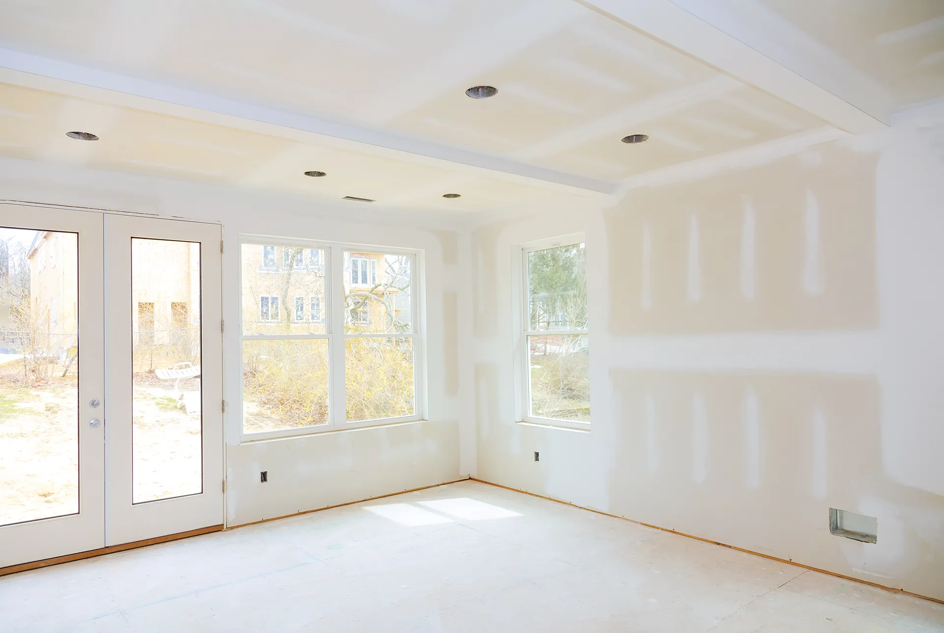 Room interior under construction with new drywall, mudded seams, recessed lights, and large windows