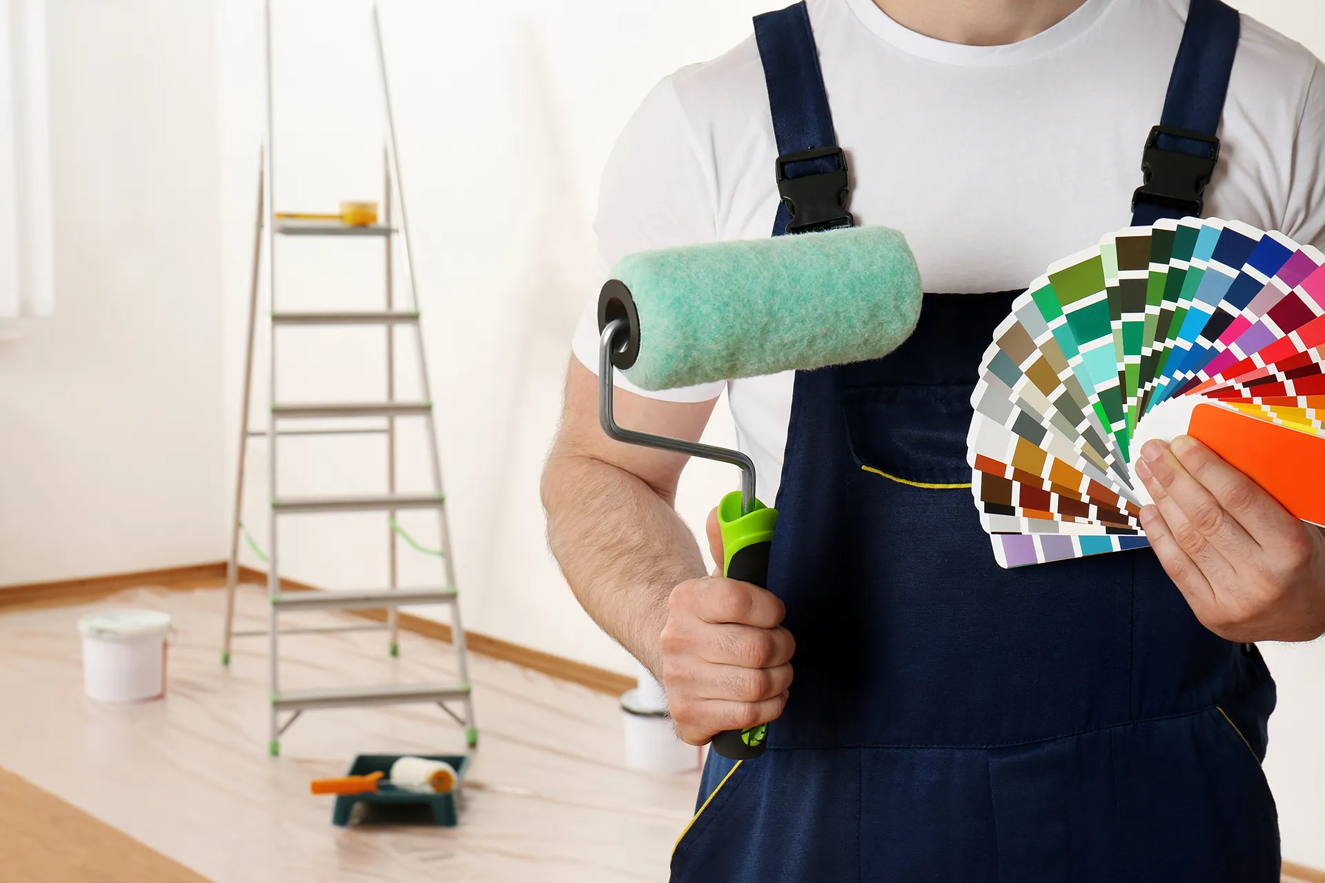 Professional house painter in overalls holding a paint roller and a color swatch fan deck for wall renovation